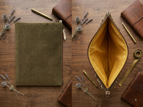 A split view of an olive green corduroy zippered pouch on a wooden tabletop, with one side closed and the other side open to show a mustard yellow interior. Styled with leather notebooks, brass pens, and dried lavender sprigs.