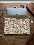 A floral laptop sleeve with an olive green corduroy top, shown holding a gray laptop on a wooden desk with styled props including a leather journal, pen, and dried lavender.