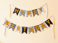 A two-tiered fabric banner garland spelling out 'HAPPY BIRTHDAY' in mustard yellow letters, decorated with navy blue, white, and blue patterned pennant flags, hanging against a light beige wall.