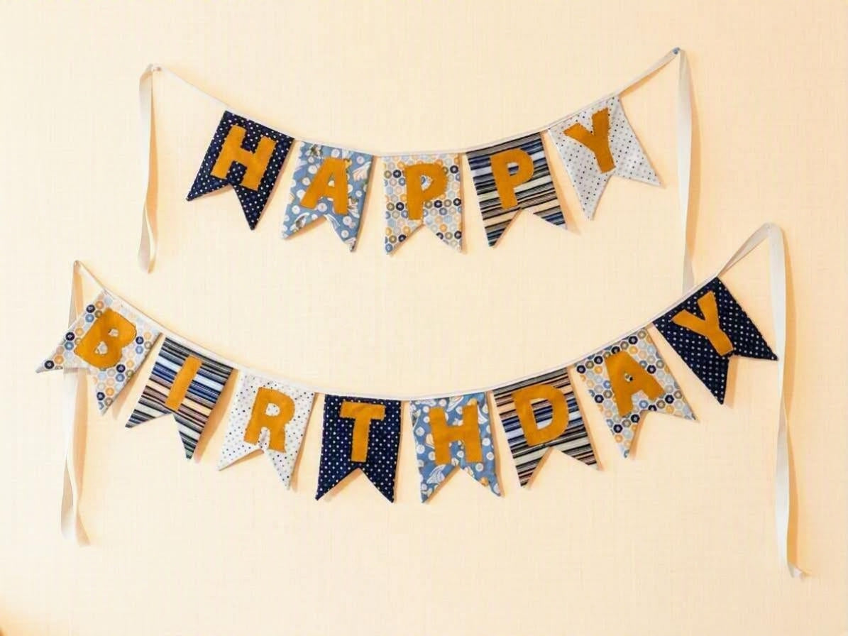 A two-tiered fabric banner garland spelling out 'HAPPY BIRTHDAY' in mustard yellow letters, decorated with navy blue, white, and blue patterned pennant flags, hanging against a light beige wall.