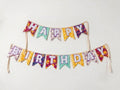 A colorful 'Happy Birthday' banner made of triangular flags with floral and patterned designs in purple, yellow, teal, and pink, hung in two rows against a plain white wall.