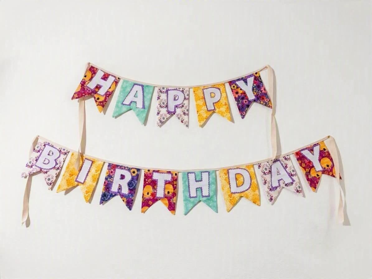 A colorful 'Happy Birthday' banner made of triangular flags with floral and patterned designs in purple, yellow, teal, and pink, hung in two rows against a plain white wall.