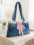 Pink Bow Denim Upcycled Tote featured in Blue Jean Purse.PNG showcasing a shoulder bag handcrafted from the waistline of repurposed blue jeans, detailed with a pink and white gingham bow woven through the belt loops on a light wood table.