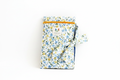 The Meadow Flora Quilted Book Sleeve & Cover featuring a blue and yellow botanical print on cream fabric, designed with vertical channel quilting, a gold top zipper with a matching fabric pull-tab, a side hand strap, and a centered white KIALIYAH label.