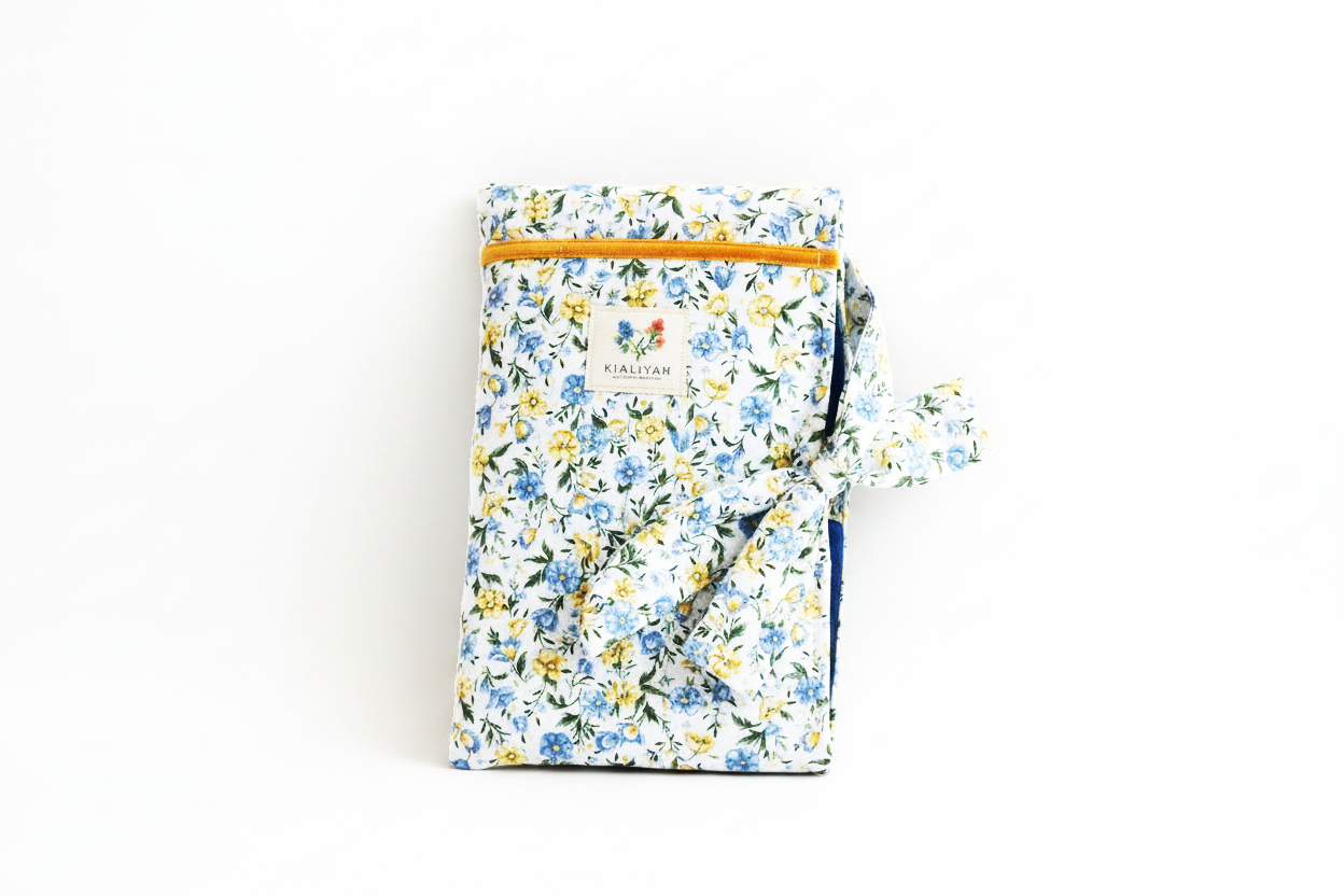 The Meadow Flora Quilted Book Sleeve & Cover featuring a blue and yellow botanical print on cream fabric, designed with vertical channel quilting, a gold top zipper with a matching fabric pull-tab, a side hand strap, and a centered white KIALIYAH label.