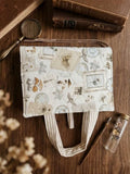 A fabric tote bag with a vintage collage print of butterflies, frames, and botanical motifs, styled on a wooden tabletop with books, a magnifying glass, and dried flowers.
