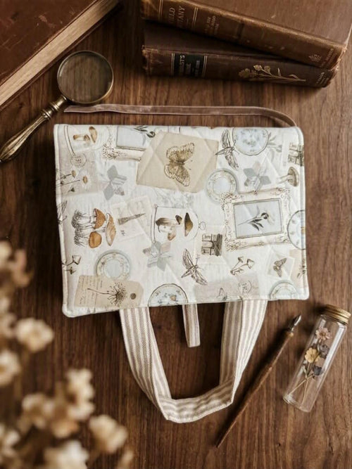 A fabric tote bag with a vintage collage print of butterflies, frames, and botanical motifs, styled on a wooden tabletop with books, a magnifying glass, and dried flowers.