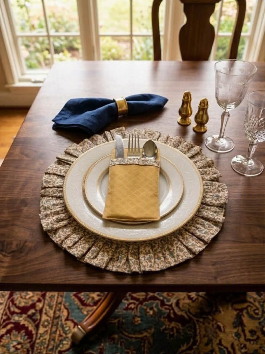 A styled dining table setting featuring a round beige and brown floral-patterned ruffled placemat under a layered white and gold charger plate, with a gold napkin, silverware, a blue napkin, and clear wine glasses on a dark wood table.