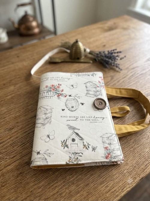 A small fabric journal or notebook with a cream-colored cover, printed with delicate black line drawings of bees, flowers, and birds, and a brown button closure. It is resting on a wooden table with a yellow strap and dried lavender in the background.