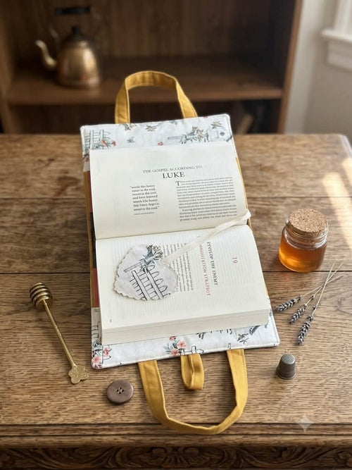 An open book with printed text on the pages, wrapped in a floral-patterned fabric cover with yellow fabric handles, styled on a rustic wooden table with small decorative props including a honey dipper, a cork bottle, and dried lavender.