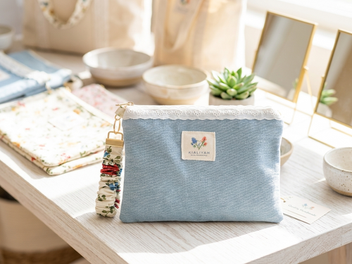 Country Meadow Upcycled Denim showcasing a light blue denim zippered pouch with white eyelet lace trim and a floral fabric scrunchie wristlet strap displayed on a white wooden table.
