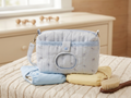 Blue Elephant Quilted Diaper Pouch featured in Diaper-Wipe Pouch.png showcasing a light blue mini elephant print fabric with geometric trim, a center wipe dispenser window, and a snap closure flap resting on cloth diapers and a knit blanket.