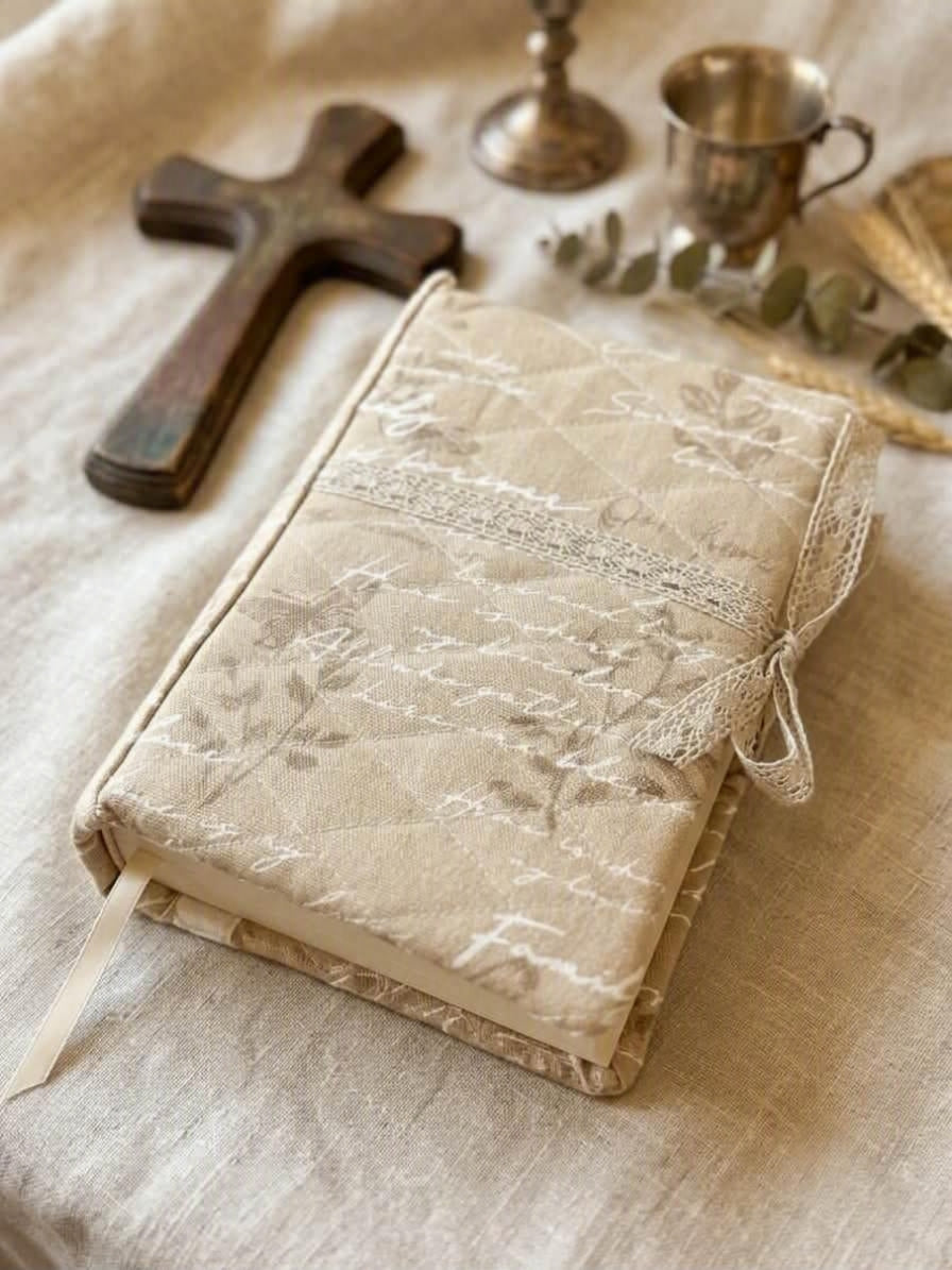 Heirloom Script Quilted Premium Bookcover showcasing a plush oatmeal linen book jacket with cursive handwriting, diamond quilting, and delicate lace ribbon trim resting on a limestone slab with dried flowers.
