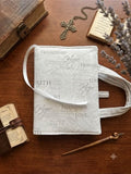 A white fabric Bible cover with embossed script words like 'Friendship', 'Hope', 'Faith', and 'Love', styled on a wooden tabletop with a cross necklace, old book, notebook, and dried lavender.