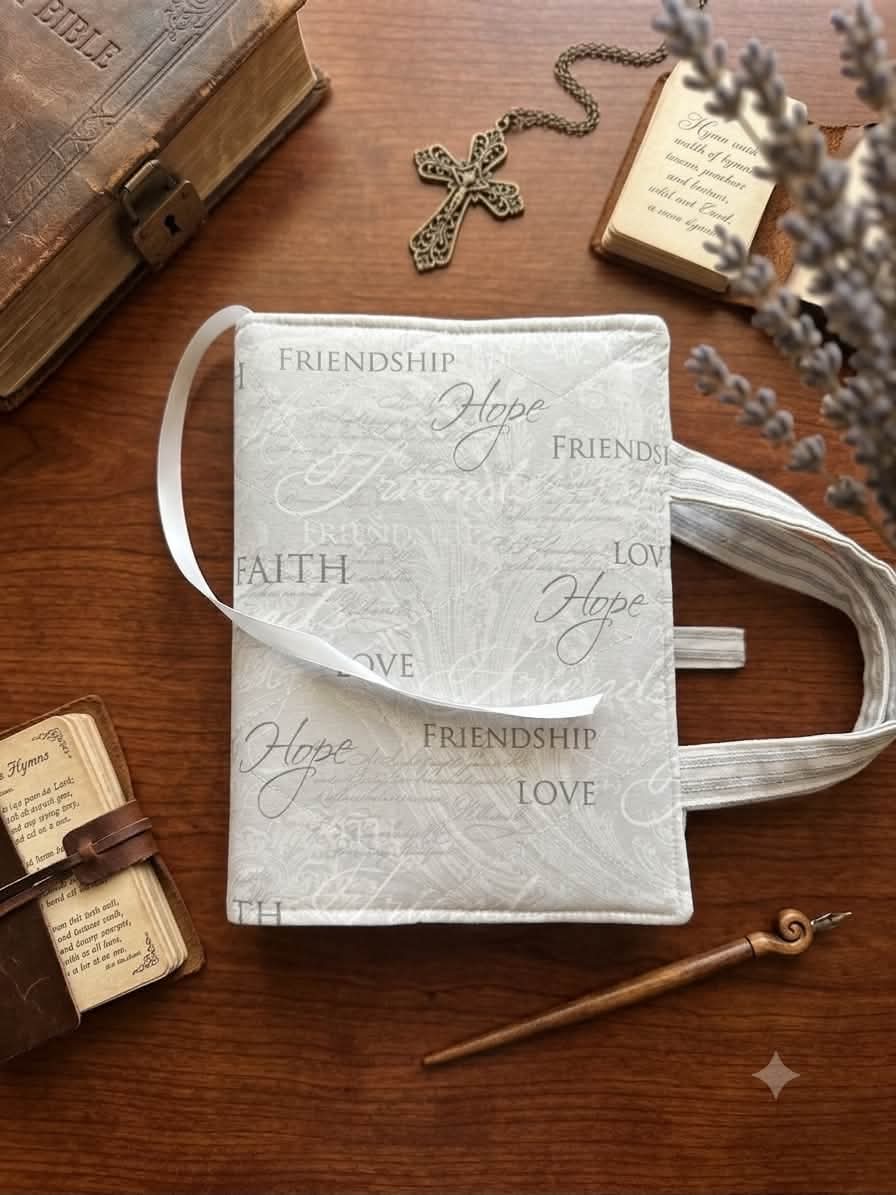 A white fabric Bible cover with embossed script words like 'Friendship', 'Hope', 'Faith', and 'Love', styled on a wooden tabletop with a cross necklace, old book, notebook, and dried lavender.