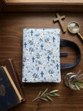 A quilted fabric Bible cover with a light blue background, navy blue crosses, and white floral motifs, displayed on a dark wooden table with decorative props including a magnifying glass, a wooden cross, a book, and a small plant.