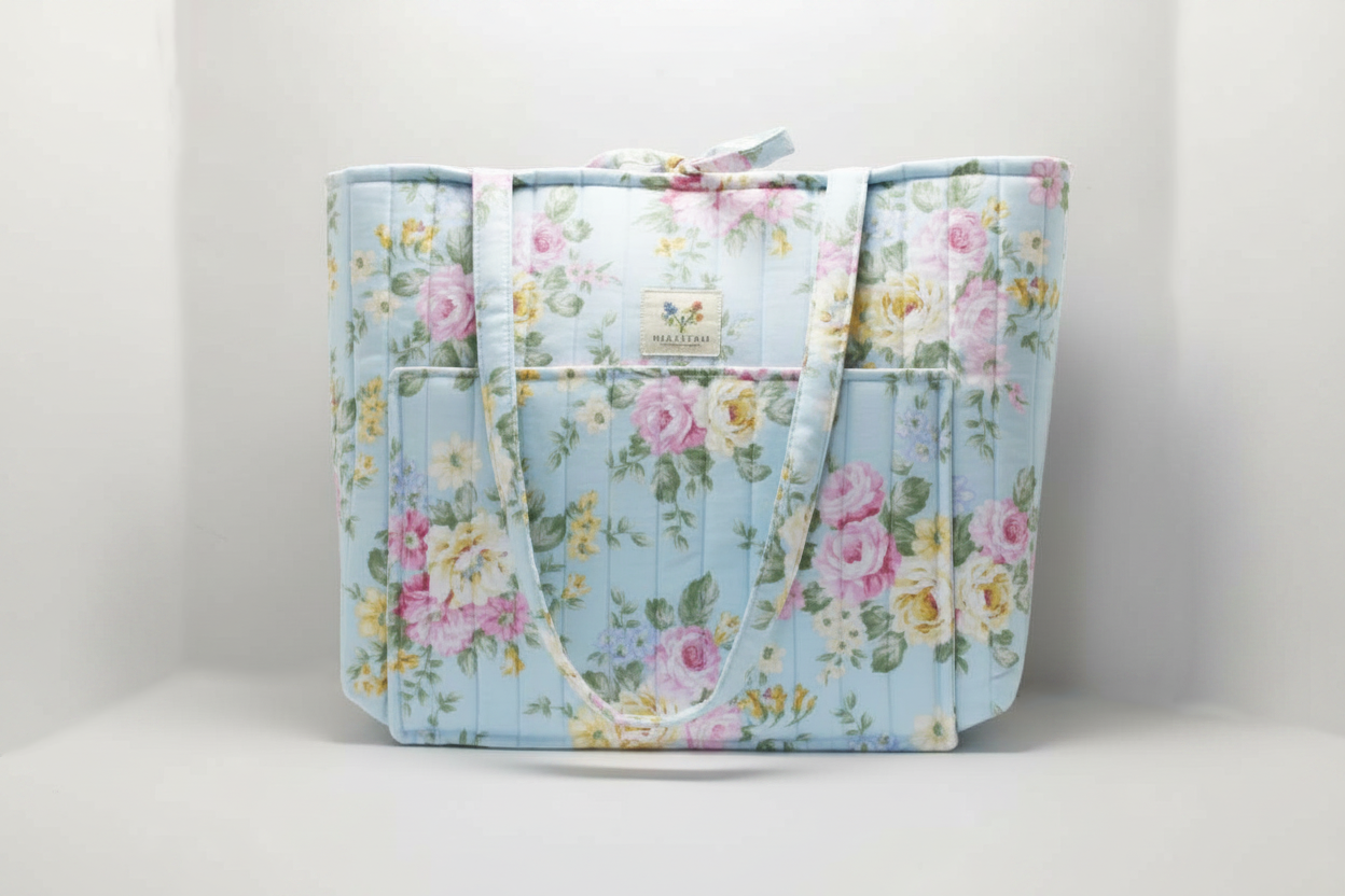 The English Rose Quilted Market Tote featuring a vintage pink and yellow rose print on sky-blue cotton fabric, designed with vertical channel quilting, a front slip pocket, and matching fabric shoulder straps.