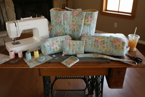 English Rose Quilted Travel Bundle featuring a matching vertical-quilted light blue canvas tote bag, two cosmetic pouches, an accessory case, an ID wallet, and a wristlet strap on a wooden sewing table next to a sewing machine.