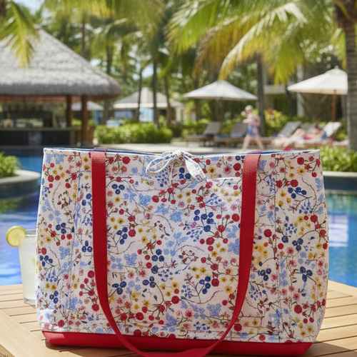 The Crimson Botanical Quilted Resort Tote featuring a vibrant cherry red, blue, and yellow floral print, a saturated red canvas base, and red shoulder straps, sitting poolside on a sunny day.