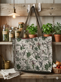 French Market Herb Quilted Carryall canvas grocery bag featuring a cream botanical print with rosemary and parsley, wide olive green shoulder straps, and a central fabric brand label on a wooden shelf.