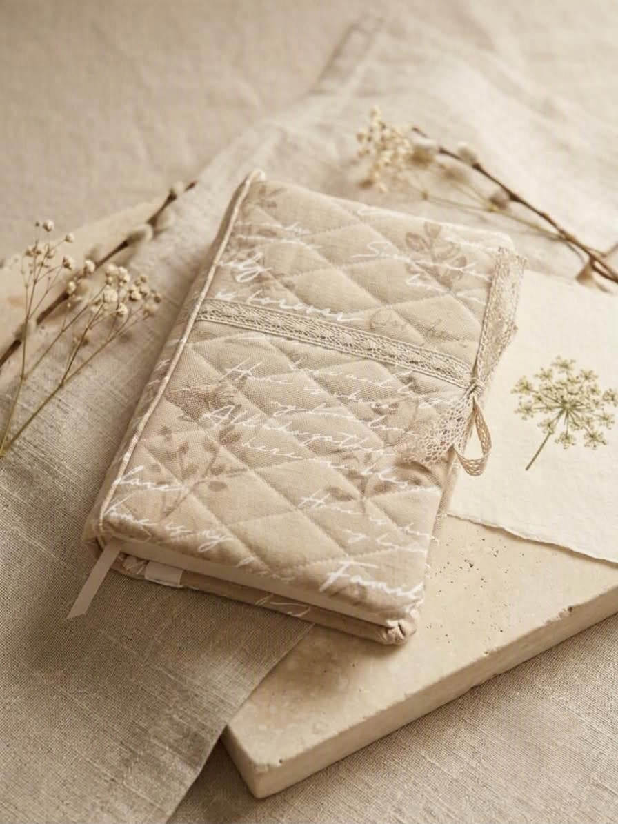 Heirloom Script Quilted Premium Bookcover showcasing a plush oatmeal linen book jacket with cursive handwriting, diamond quilting, and delicate lace ribbon trim resting on a limestone slab with dried flowers.