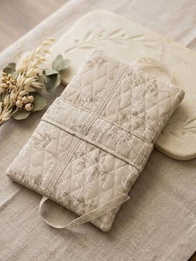 A quilted cream-colored fabric pouch with a diamond pattern and a matching elastic strap, styled on a linen surface with dried flowers and a decorative stone plate.