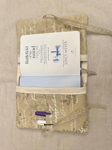 A beige fabric notebook pouch with a script pattern, holding a white notebook, a light blue book, a purple pen, and a white highlighter, laid flat on a textured beige fabric background.