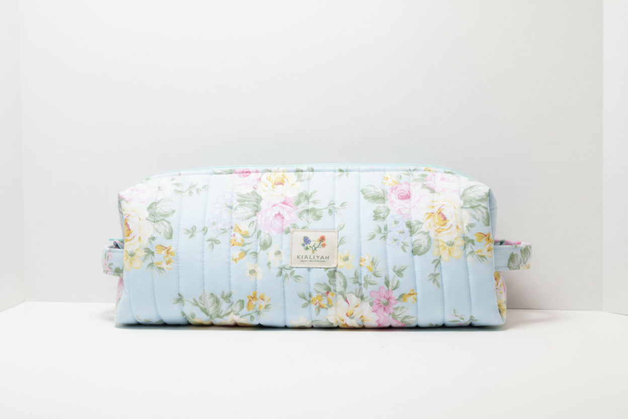 The English Rose Quilted Hot Tool & Travel Bag featuring a vintage pink and yellow floral print on sky-blue cotton fabric, designed with vertical channel quilting, a gold top zipper with a matching fabric pull-tab, a side hand strap, and a centered white KIALIYAH label.