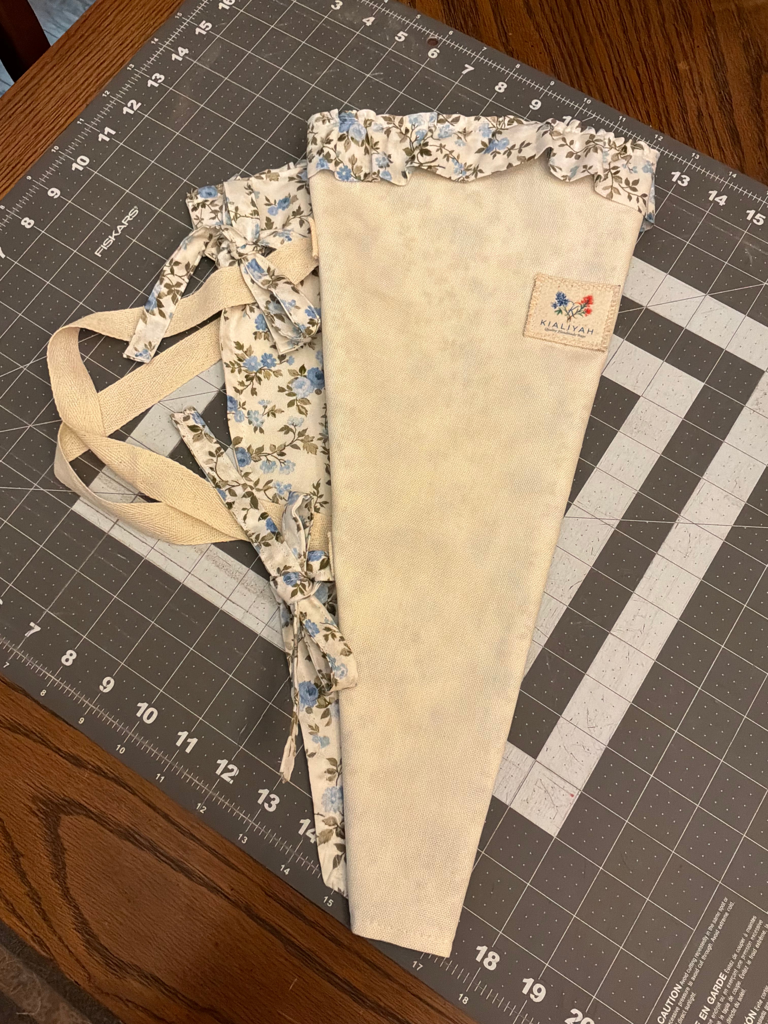 Wimberley Mist and Blossom Bouquet Wrap canvas flower carrier featuring a textured cream canvas with light blue floral ruffles and matching fabric side ties on a gray cutting mat.