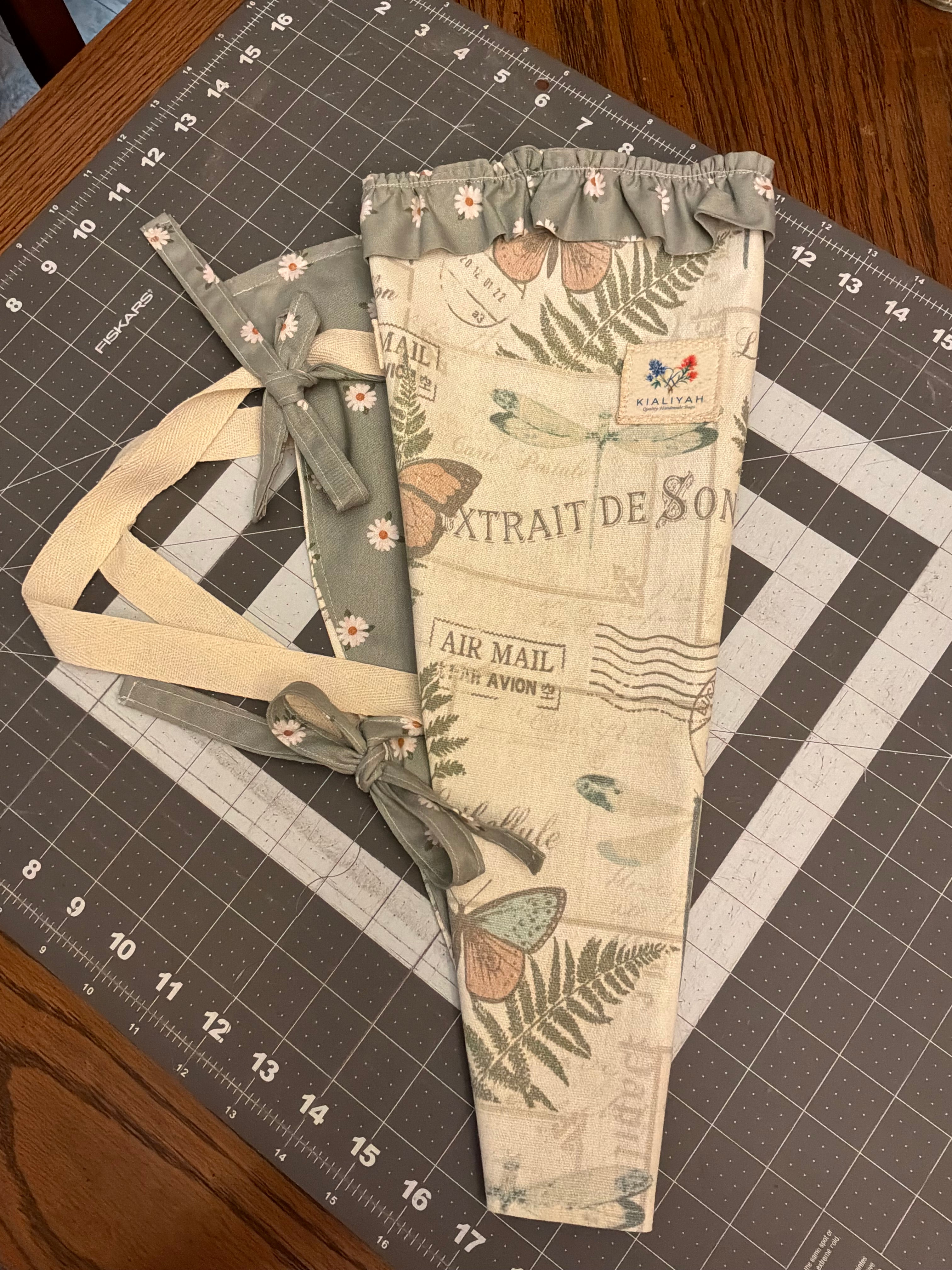 Post Oak Heritage Bouquet Wrap canvas flower carrier featuring a vintage cream botanical script fabric with butterflies and ferns, paired with a muted sage green daisy ruffle top and matching side ties on a gray cutting mat.