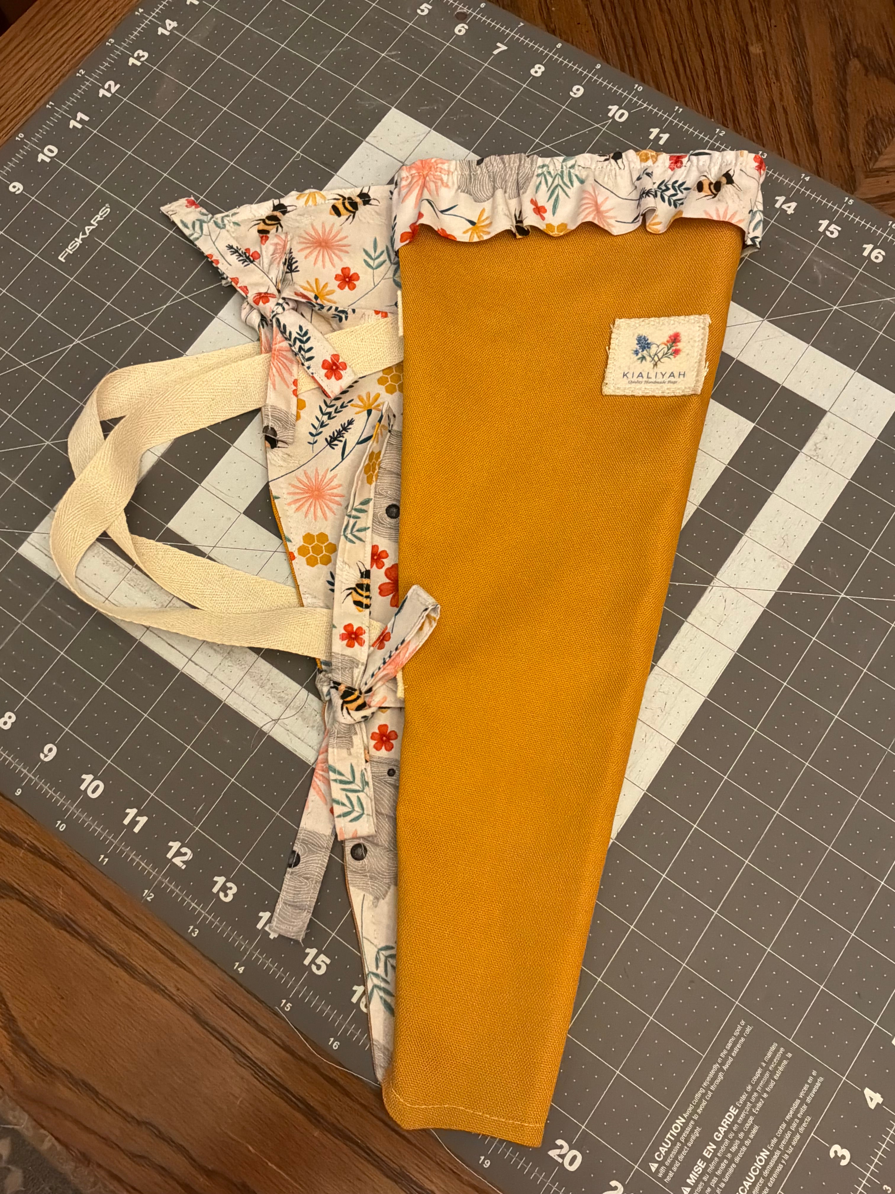 Cibolo Creek Honey and Blossom Bouquet Wrap canvas flower carrier featuring a vibrant marigold gold canvas with cream honeybee and wildflower ruffles and matching fabric side ties on a gray cutting mat.