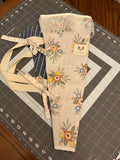 Gruene Lace and Indigo Bouquet Wrap canvas flower carrier featuring a cream canvas with ochre and peach floral prints, a detailed scalloped cream lace trim at the top opening, and matching fabric side ties on a gray cutting mat.