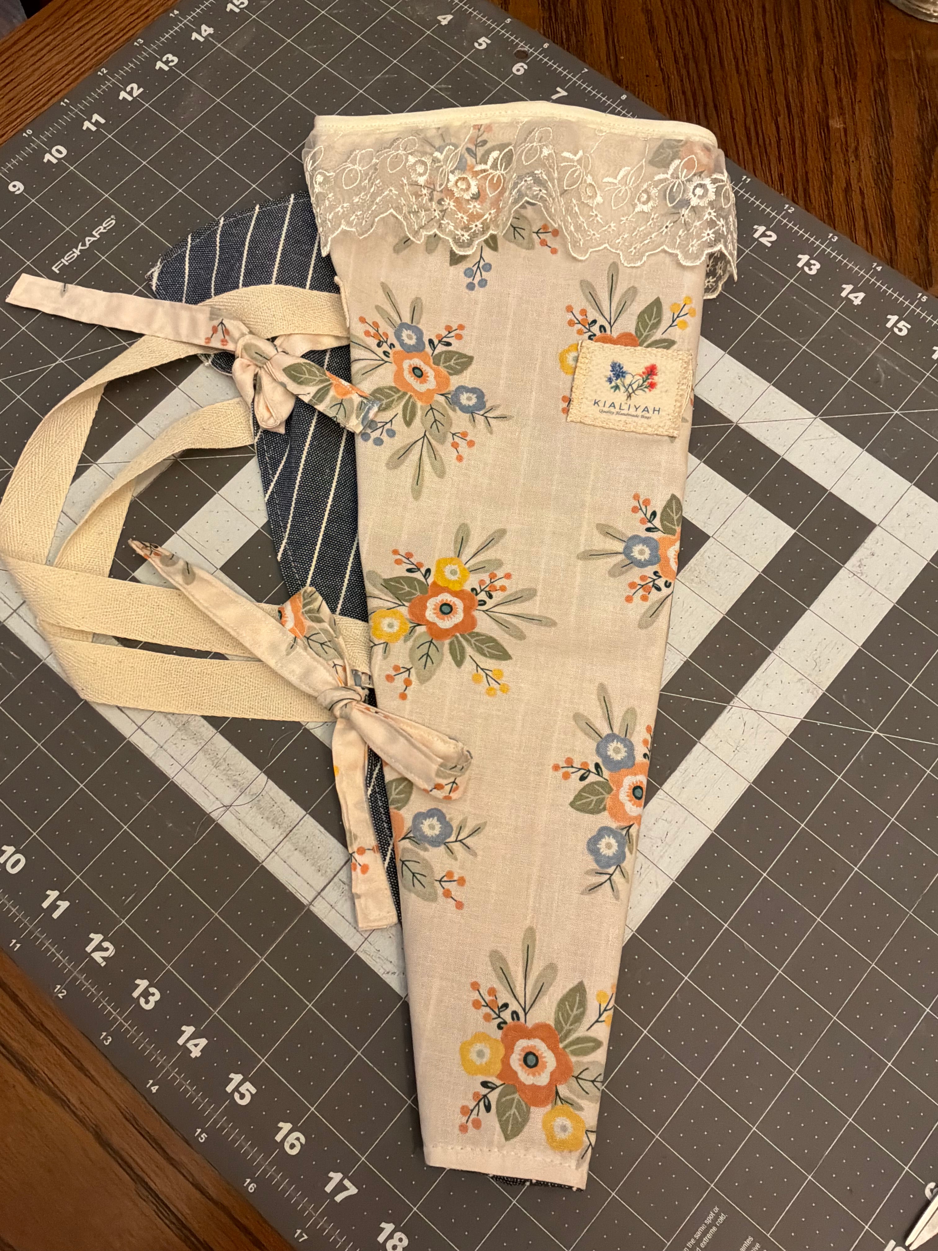 Gruene Lace and Indigo Bouquet Wrap canvas flower carrier featuring a cream canvas with ochre and peach floral prints, a detailed scalloped cream lace trim at the top opening, and matching fabric side ties on a gray cutting mat.