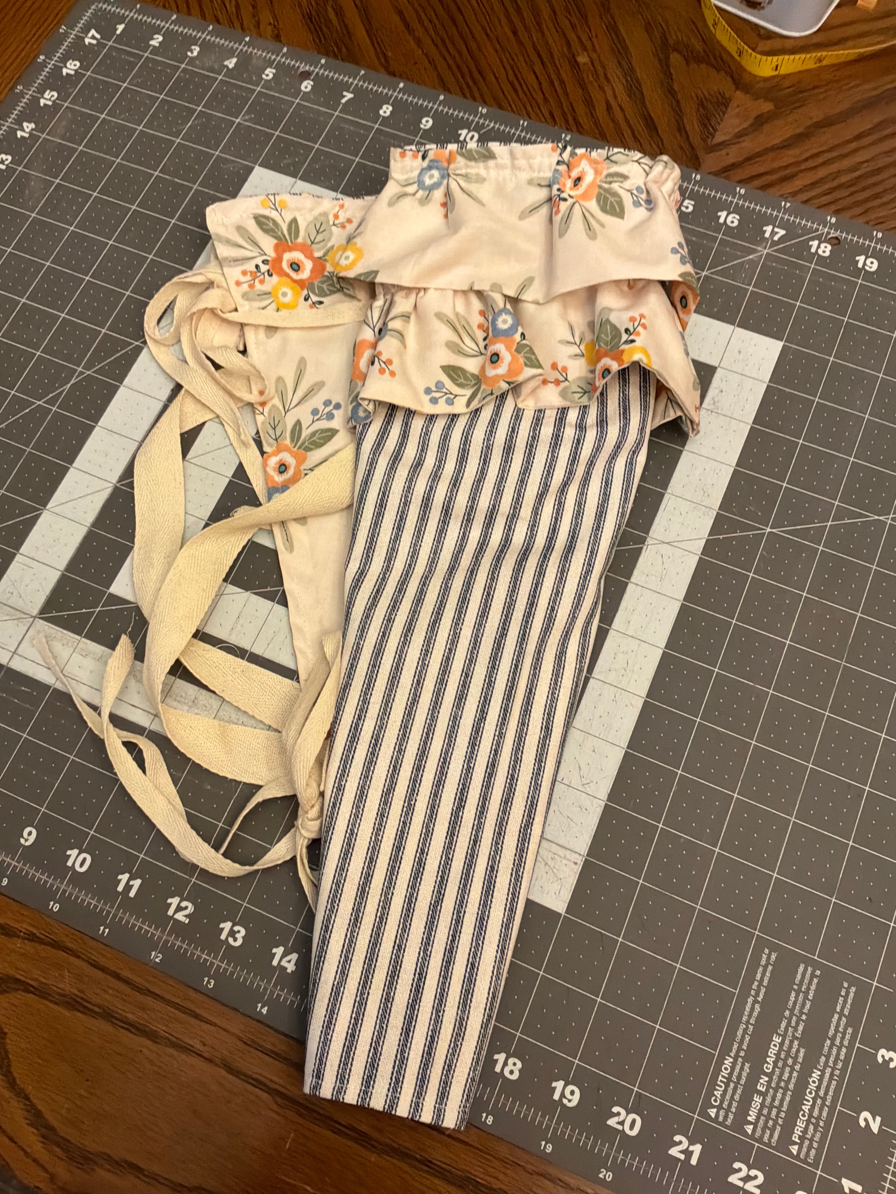Concan Double-Ruffle and Stripe Bouquet Wrap canvas flower carrier featuring a classic navy and cream ticking stripe canvas base with a double layered ochre and peach floral print ruffle top and matching side ties on a gray cutting mat.