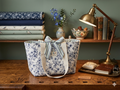 The Blue Toile & Mixed Stripe Quilted Market Tote featuring a classic blue pastoral toile top print, a blue and white ticking stripe quilted base, cream webbing shoulder straps, and matching striped top ties, resting upright on a rustic wooden workshop table.