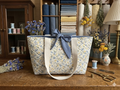 The Meadow Flora Quilted Market Tote featuring a delicate blue and yellow botanical print on cream quilted fabric, designed with wide cream webbing shoulder handles and a contrasting slate blue fabric bow tied at the top opening, resting on a rustic wooden work table.