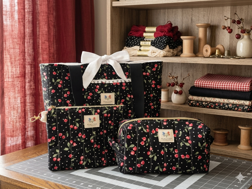 Black Cherry Jubilee Quilted Travel Bundle featuring a matching black canvas tote bag with a white bow, a boxy cosmetic case, and a flat zipper wallet pouch with a cherry and polka dot print on a cutting mat.