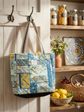 Tuscan Orchard Quilted Carryall canvas grocery bag featuring a yellow lemon and blue majolica tile patchwork print, flat cream shoulder straps with blue trim, and a central brand label hanging from a hook.
