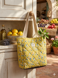 Capri Orchard Quilted Carryall canvas grocery bag featuring a mustard yellow print with lemons and white flowers, wide cream shoulder straps, and a central brand label hanging from a white cabinet hook.