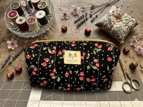 Black Cherry Jubilee Quilted Makeup Bag boxy cosmetic case featuring a black and white polka dot cherry print with vertical stitching and a gold zipper closure on a wooden workspace.