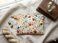 Gold Blossom Quilted Wallet clutch featuring a vibrant multi-colored wildflower print, structured diamond stitching, a gold top zipper with a golden-yellow fabric pull tab, and an antique brass swivel clasp on a cream linen cloth next to vintage books.
