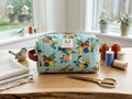 Terracotta Songbird Quilted Makeup Bag showcasing a detailed songbird print on an aqua blue background, vertical channel quilting, a rich rust orange side handle, and a top zipper on a light wooden table.