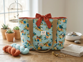 Terracotta Songbird Quilted Tote bag featuring a detailed songbird print on an aqua blue background, a rich rust orange top bow closure, and khaki canvas shoulder straps on a light wooden workshop table.