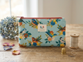 Terracotta Songbird Quilted Wallet clutch featuring a detailed wildbird print on an aqua blue background, a secure rust orange top zipper, and a central brand label on a wooden table next to a sewing thimble and thread spool.