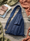 Navy Teardrop Sling Bag showcasing a navy blue canvas crossbody bag with multiple zippered pockets, a snap flap compartment, and a cross-stitch patterned strap resting on a rustic wood boardwalk beside a leather notebook and maps.