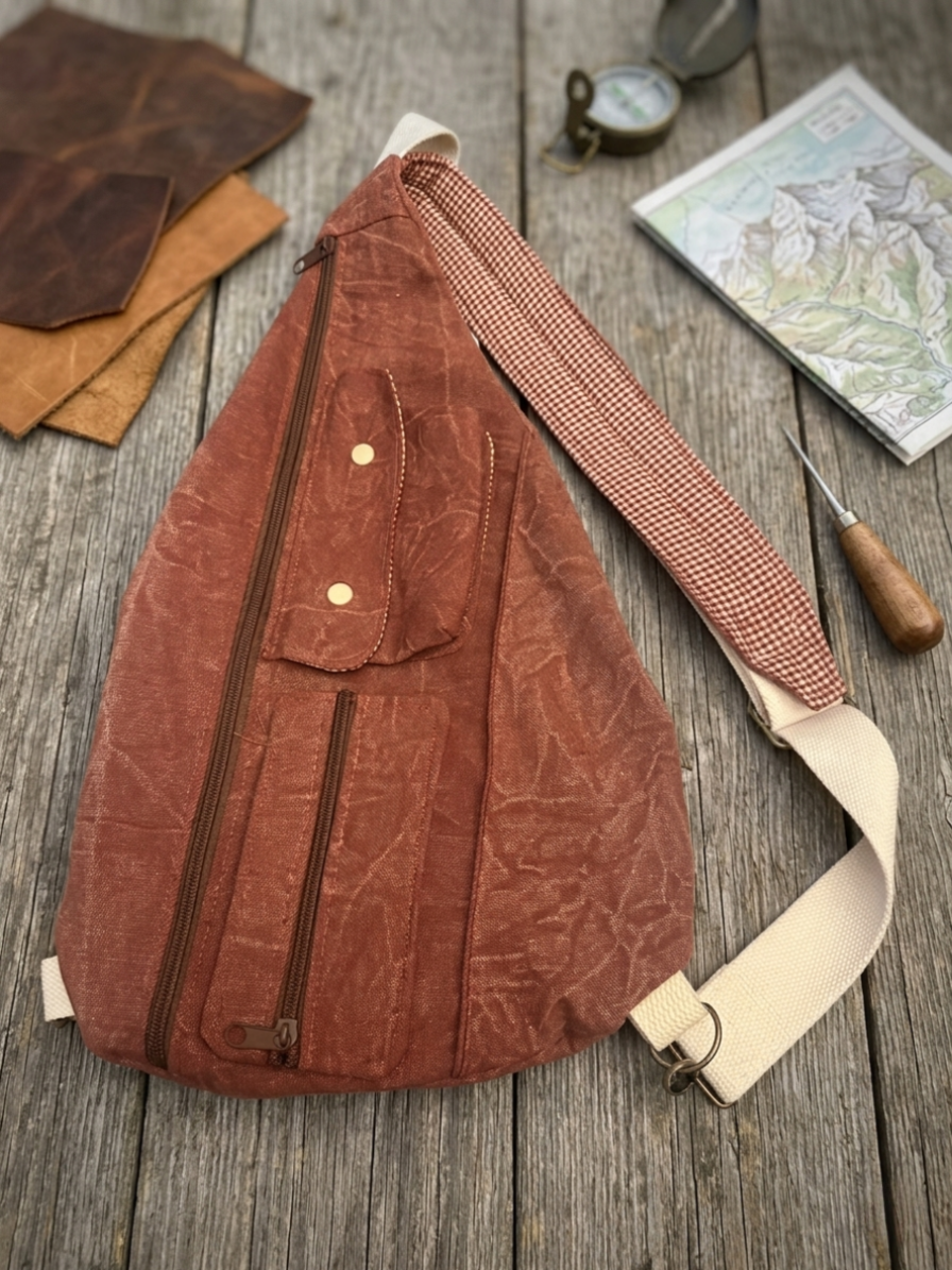 Rust Teardrop Sling Bag showcasing a terracotta canvas crossbody bag with multiple zippered pockets, a snap flap compartment, and a cream and burgundy gingham checkered patterned strap resting on a grey Fiskars cutting mat.