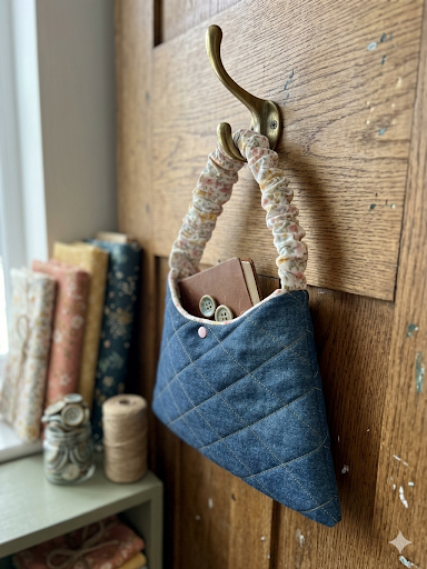 Blue fabric basket with floral handle hanging on a wooden wall.