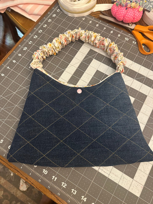 Denim Scrunchie Purse showcasing a dark indigo diamond-quilted denim handbag with gold topstitching, a pink center snap, and a gathered floral fabric scrunchie handle resting on a grey Fiskars cutting mat.
