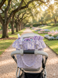 Purple Daisy Quilted Stroller Bag showcasing a lavender daisy print fabric with a gathered ruffle top collar, hanging from a grey stroller handlebar on a sunlit park path lined with trees and flowers.