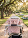 Pink Blossom Quilted Stroller Bag showcasing a pink ditsy floral print fabric with a fairytale woodland ruffle top collar, hanging from a grey stroller handlebar on a sunlit park path lined with trees and flowers.